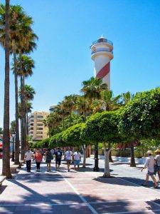 People walking on a sunny promenade lined with palm trees and a tall red and white striped lighthouse